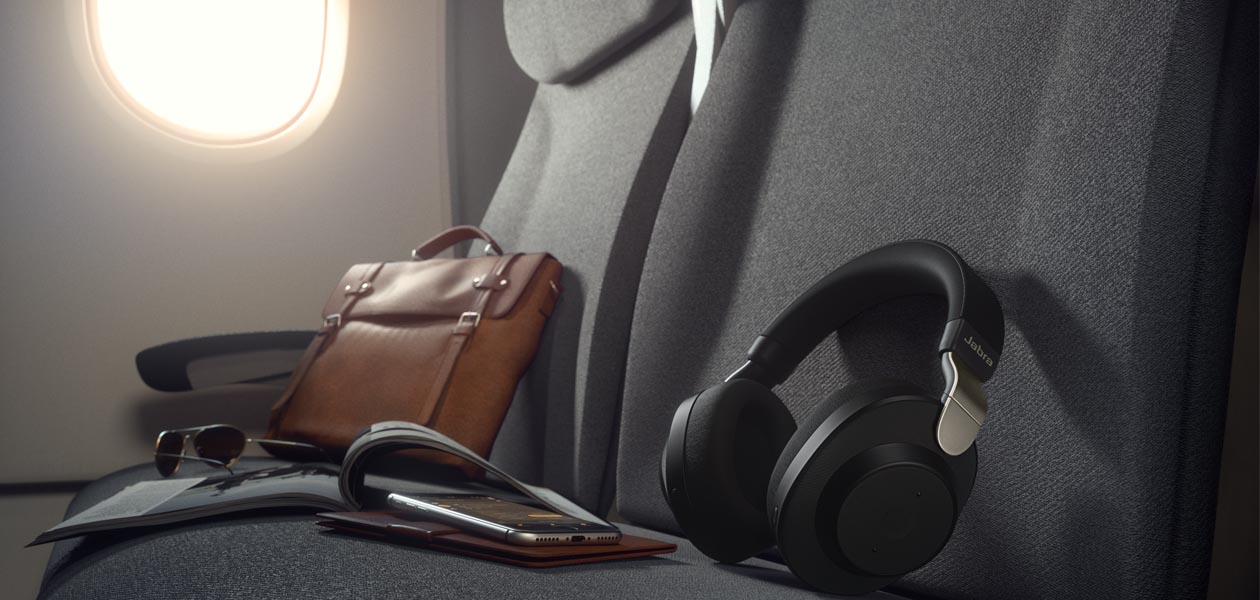 Langattomat Jabra Elite 85h around-ear-kuulokkeet Jabra Elite 85h -kuulokkeet lentokoneen istuimella puhelimen ja salkun vieressä.