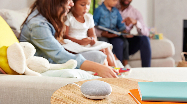 Alle familimedlemmer kan legge til produkter på handlelisten En Google Home Mini på et bord ved siden av en familie i en sofa