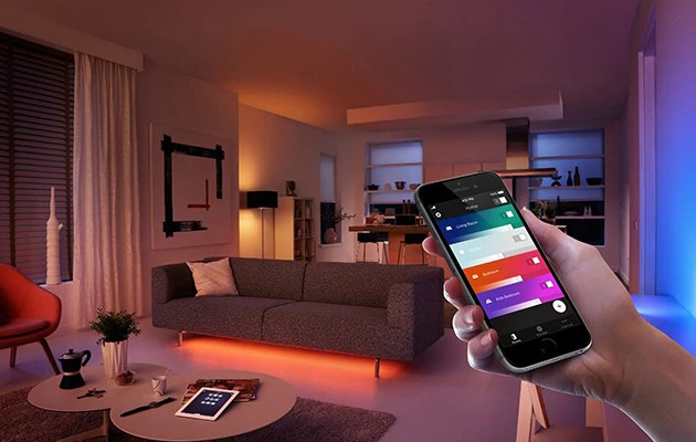 Synkroniser belysningen med soloppgang og solnedgang Hånd som holder smarttelefon foran fargerikt opplyst stue.