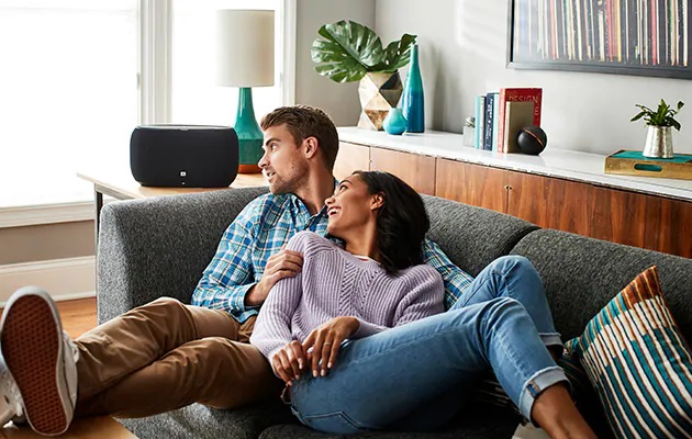 JBL Link høyttalere fungerer med Google Assistant En kvinne og en mann slapper av på sofaen. En stor høyttaler står på bordet ved siden av.