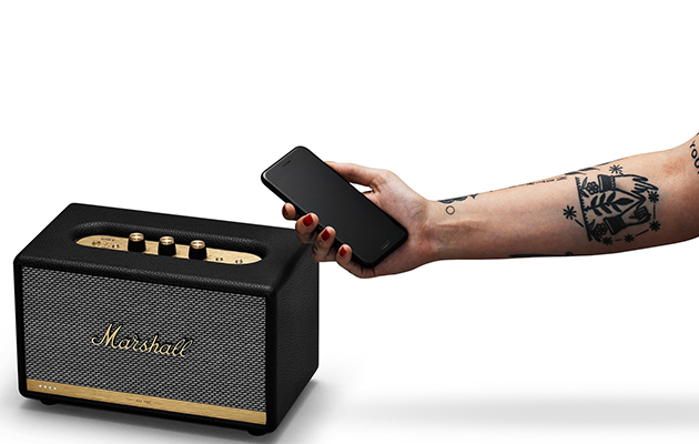 En annen smarthøyttaler med Google Assistant er Marshall Acton En tattovert hånd holder en mobiltelefon ved siden av en høyttaler i sort og gull med Marshall-logo.