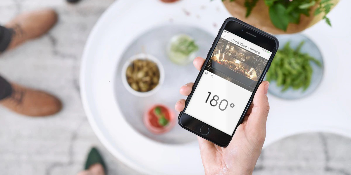 Hvorfor er en stekeovn med WiFi en god idé? En hånd som holder en mobil som viser temperatur og direktevideo fra innsiden av stekeovnen.