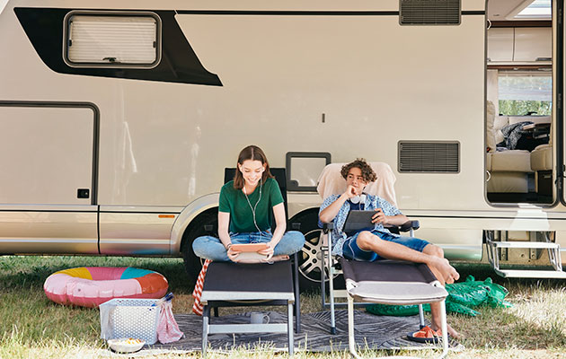 Underholdning på ferie To tenåringer, en gutt og en jente, sitter i campingstoler foran en bobil og kikker på nettbrett