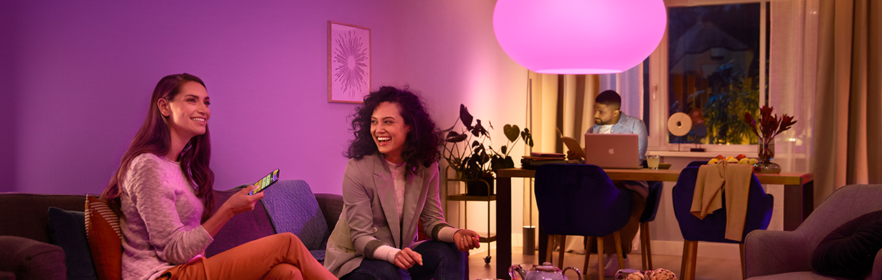 Philips Hue – Bridge vs Bluetooth To kvinner sitter i en sofa og en mann sitter ved et bord i bakgrunnen.
