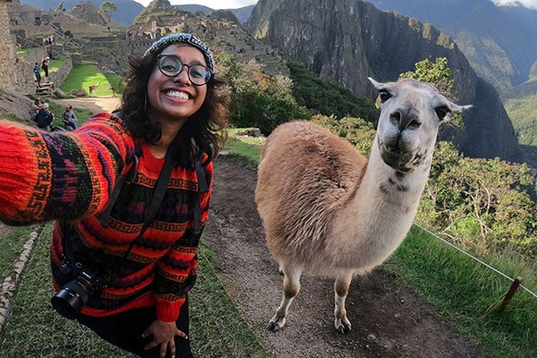 En person tar en selfie sammen med en llama, søramerikanske fjell i bakgrunnen