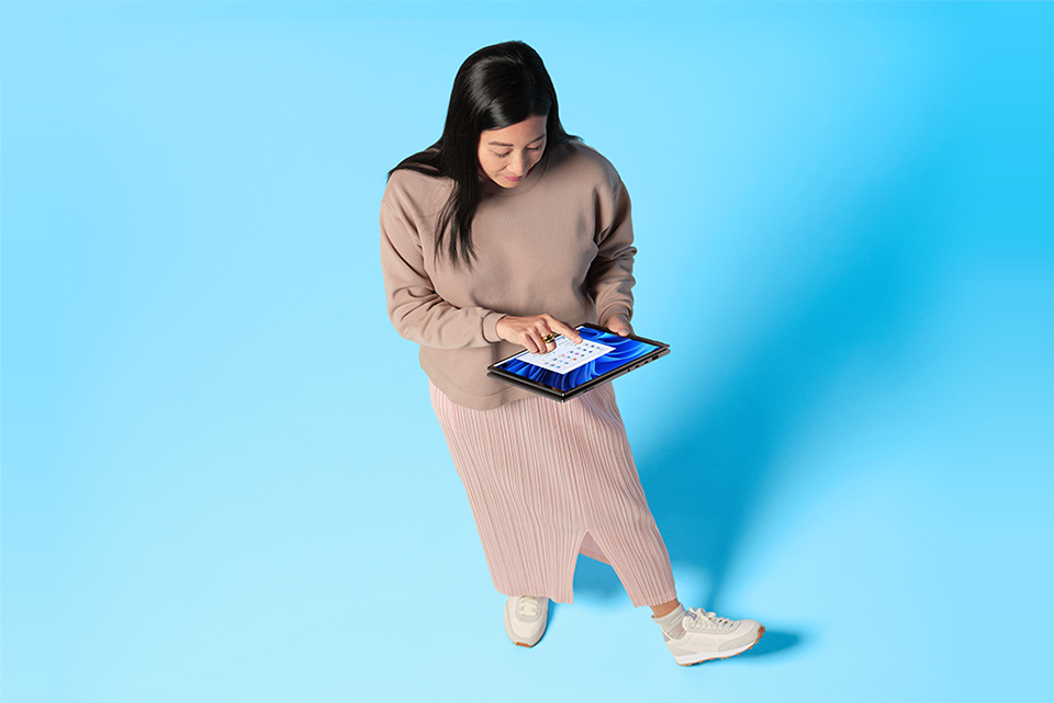 Få et nytt perspektiv Kvinne med beige klær står med Windows 11 2-i-1 i hendene foran en blå bakgrunn