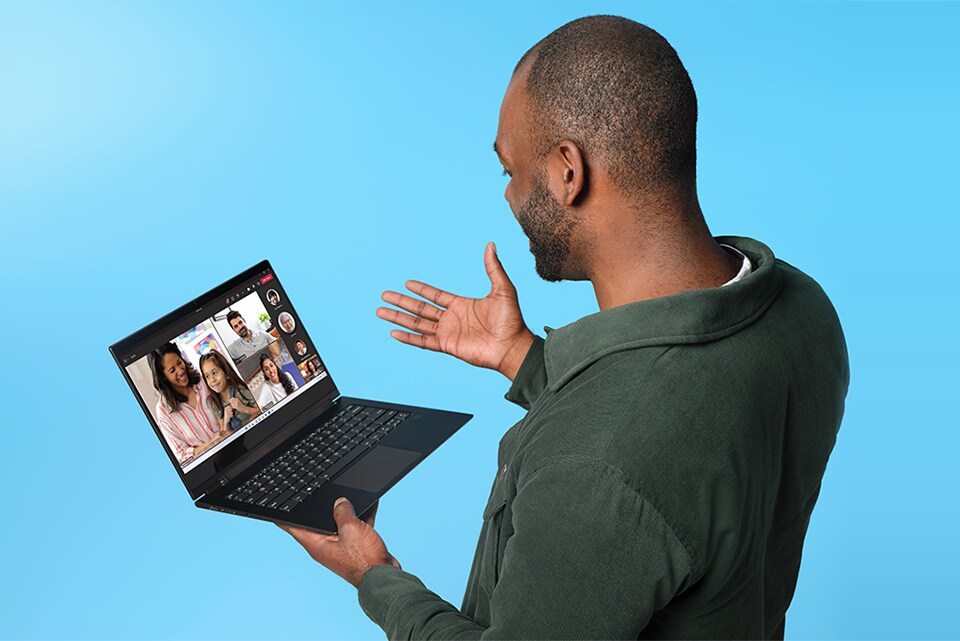 Mann med mørkegrønn jakke holder en liten Windows 11-laptop i hånden og prater med folk på Teams