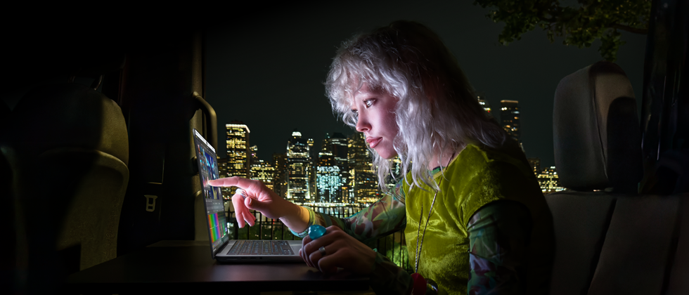 En person sitter i baksetet i en van, ved den åpne passasjerdøren er det visning av en byskygge om kvelden. En MacBook Pro med PROLED XDR-skjerm står på et arbeidsbord. Personen er opplyst av skjermen mens vedkommende bruker Touch-funksjonen til å redigere en video i DaVinci Resolve Studio