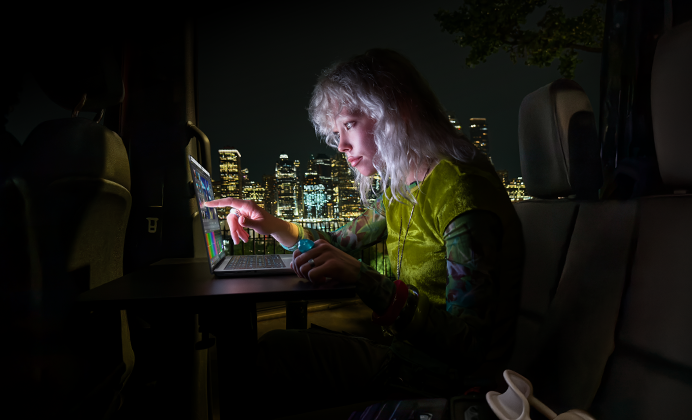 En person sitter i baksetet i en van, ved den åpne passasjerdøren er det visning av en byskygge om kvelden. En MacBook Pro med PROLED XDR-skjerm står på et arbeidsbord. Personen er opplyst av skjermen mens vedkommende bruker Touch-funksjonen til å redigere en video i DaVinci Resolve Studio