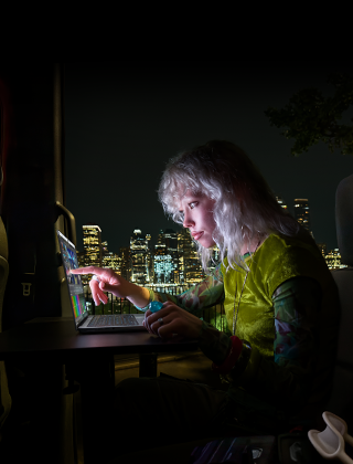 En person sitter i baksetet i en van, ved den åpne passasjerdøren er det visning av en byskygge om kvelden. En MacBook Pro med PROLED XDR-skjerm står på et arbeidsbord. Personen er opplyst av skjermen mens vedkommende bruker Touch-funksjonen til å redigere en video i DaVinci Resolve Studio