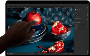 MacBook Pro med PROLED XDR-skjerm med et stillebenbilde av granatepler med blå keramikk og tekstiler, en hånd strekker seg mot skjermen for å arbeide med kunsten