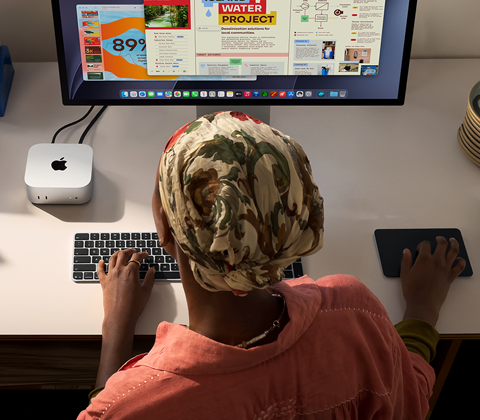 En person skriver ved et skrivebord med en Mac mini, med Studio Display tilkoblet, Magic Keyboard, Magic Trackpad og diverse objekter