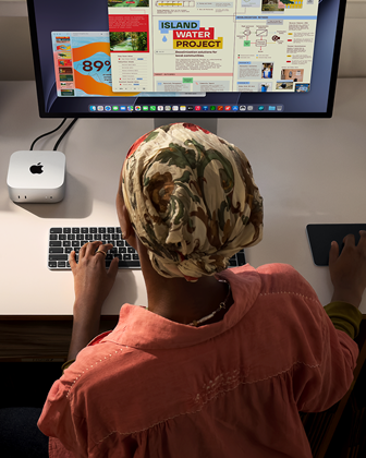 En person skriver ved et skrivebord med en Mac mini, med Studio Display tilkoblet, Magic Keyboard, Magic Trackpad og diverse objekter