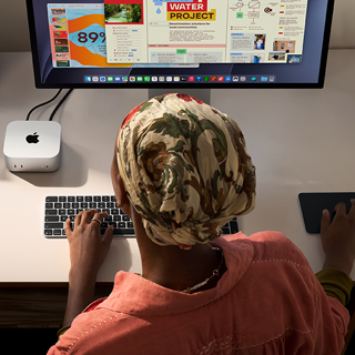 En person skriver ved et skrivebord med en Mac mini, med Studio Display tilkoblet, Magic Keyboard, Magic Trackpad og diverse objekter