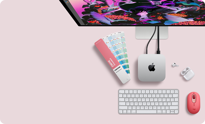 Mac mini vist ovenfra, aluminium i sølv, avrundede hjørner, blank svart Apple-logo i midten, koblet til med kabel, Studio Display er delvis synlig ovenpå. AirPods 4 er åpne, og det vises fargeprøver i Pantone samt et Logitech-tastatur og en Logitech-mus i rødt.