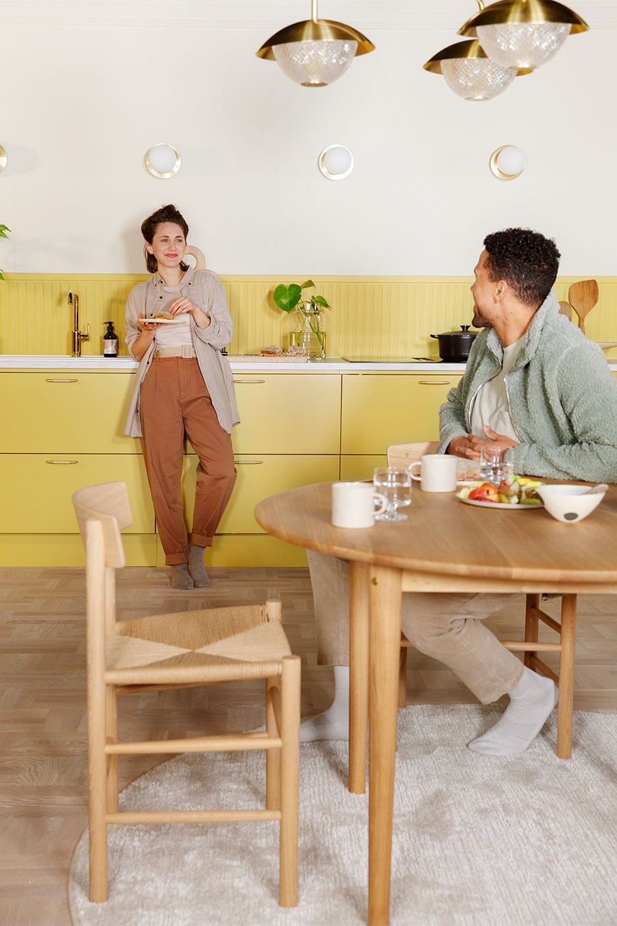 Yellow Trend Mellow kjøkken med en kvinne som spiser frokost ved vasken mens en mann sitter ved et bord og ser på henne