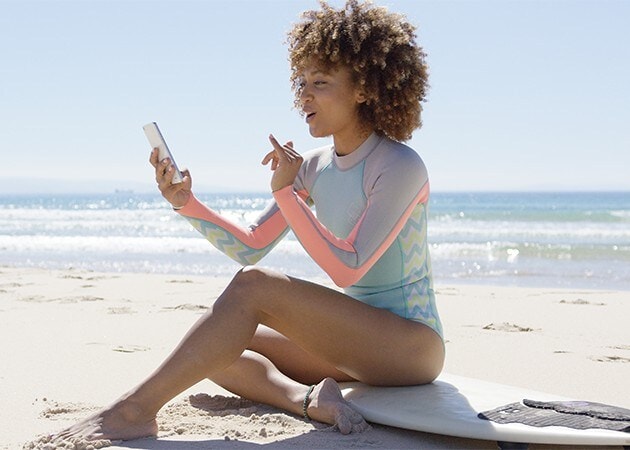 Kvinne som sitter på surfebrett på stranden mens hun bruker smarttelefon