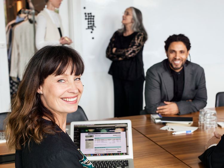 kvinne og to menn som sitter ved et konferansebord og smiler og jobber på en laptop