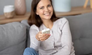 smilende kvinne sitter i sofa og holder fjernkontroll til varmepumpe