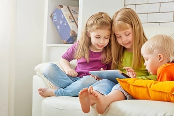 tre barn i fargerike klær bruker et nettbrett sammen i en sofa