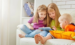 tre barn i fargerike klær bruker et nettbrett sammen i en sofa