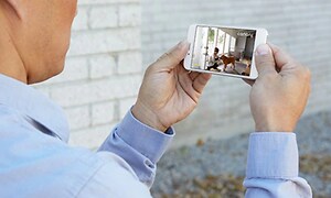 mann holder smarttelefon som viser overvåkningskamera-app med bilde fra et hus