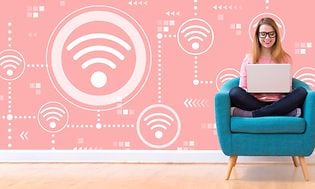 kvinne sitter i turkis stol og jobber på laptop. bak henne er en rosa vegg dekket med wifi-symboler