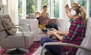 jente med hodetelefoner på ser på smarttelefon sitter i stol med gutt med nettbrett og kvinne med laptop i sofa i bakgrunnen