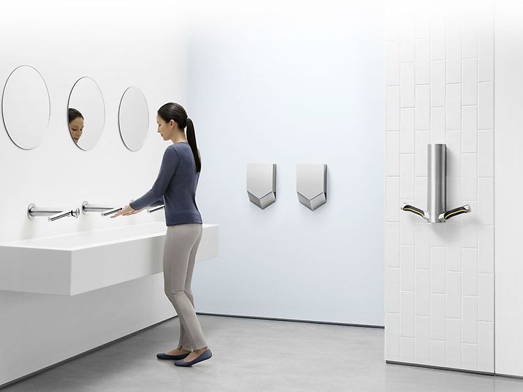 Dyson Airblade håndtørkere montert på vegg på hvitt bad hvor en kvinne vasker hendene