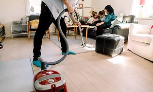 Mann som støvsuger mens familien sitter i bakgrunnen