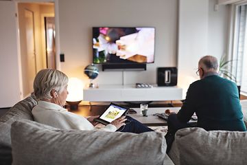 Eldre kvinne som sitter i sofa med nettbrett, mens eldre mann ser på TV i bakgrunnen