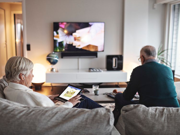 Eldre kvinne som sitter i sofa med nettbrett, mens eldre mann ser på TV i bakgrunnen