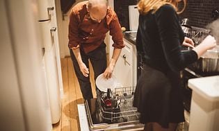 Mann og kvinne på kjøkken. Mannen putter oppvasken i oppvaskmaskinen mens kvinnen lager mat