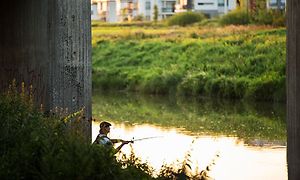 en mann sitter og fisker i en elv under en stor bro
