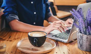 Kvinne sitter på en kafé og jobber på laptop med en kopp kaffe ved siden av