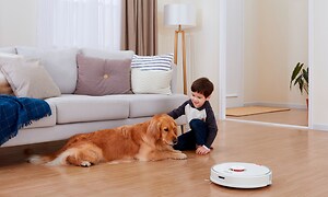 En gutt som sitter foran en sofa og klapper en hund imens en robotstøvsuger støvsuger gulvet 