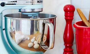 en blå KitchenAid-kjøkkenmaskin på et bord ved siden av egg og rødt kjøkkenutstyr