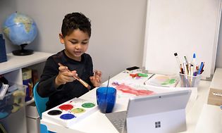 en liten gutt maler foran et bord med en bærbar Surface-datamaskin