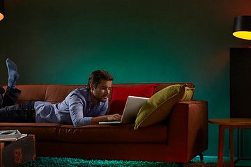 mann ligger på en sofa og bruker en bærbar pc med flere ulike lamper med smartbelysning stående rundt ham