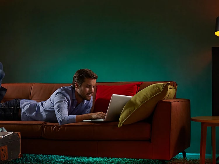 mann ligger på en sofa og bruker en bærbar pc med flere ulike lamper med smartbelysning stående rundt ham