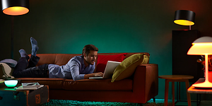 mann ligger på en sofa og bruker en bærbar pc med flere ulike lamper med smartbelysning stående rundt ham
