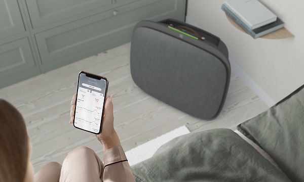 En mørkegrå Well 5 på gulvet og en kvinne med Wellbeing-appen på telefonen