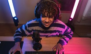 En gamer sitter foran en skjerm, med lys bak ham og en mikrofon foran.
