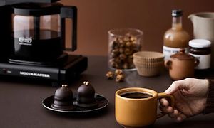 Hånd som tar kaffekopp foran tallerken med dessert og Moccamaster kaffetrakter