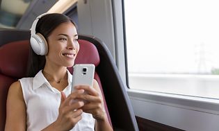 Kvinne i profesjonelt antrekk sitter på et tog med smarttelefon i hånden og støyreduserende hodetelefoner på