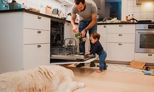 En mann og et barn legger inn skitne tallerkner i oppvaskmaskinen og en hund ligger på gulvet