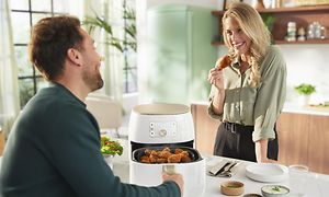 Mann som drar ut formen til en Philips Airfryer full av stekte kyllinglår og kvinne som skal til å spise et av de