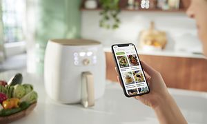 En hånd som holder en telefon med Philips NutriU-appen og en Philips Airfryer i bakgrunnen