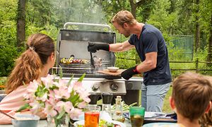 en mann griller mat på en gassgrill mens familien følger spent med