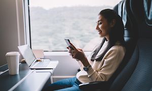 En kvinne sitter på toget og ser på smarttelefonen med en bærbar pc foran seg